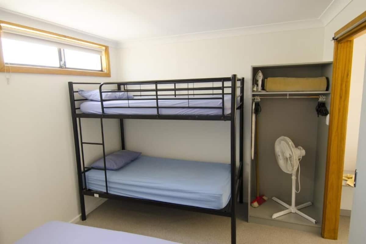 Une chambre avec un lit superposé, une fenêtre, un ventilateur et une étagère.