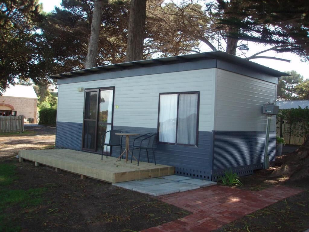 A small, single-story mobile home with a front porch, a small table and chairs, and a window.