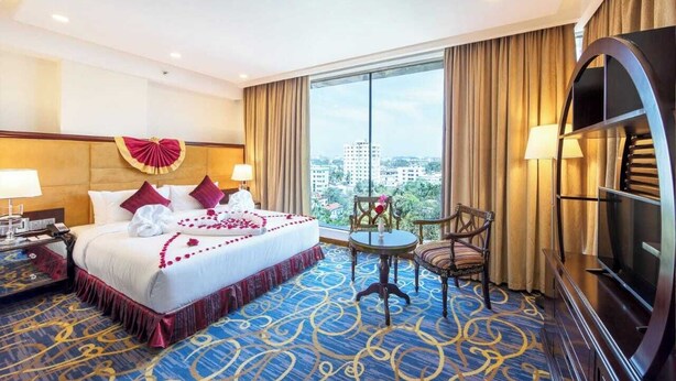 A hotel room with a large bed, a TV, a desk, and a view of the city. - Chayabithi beach resort (Cox's Bazar)