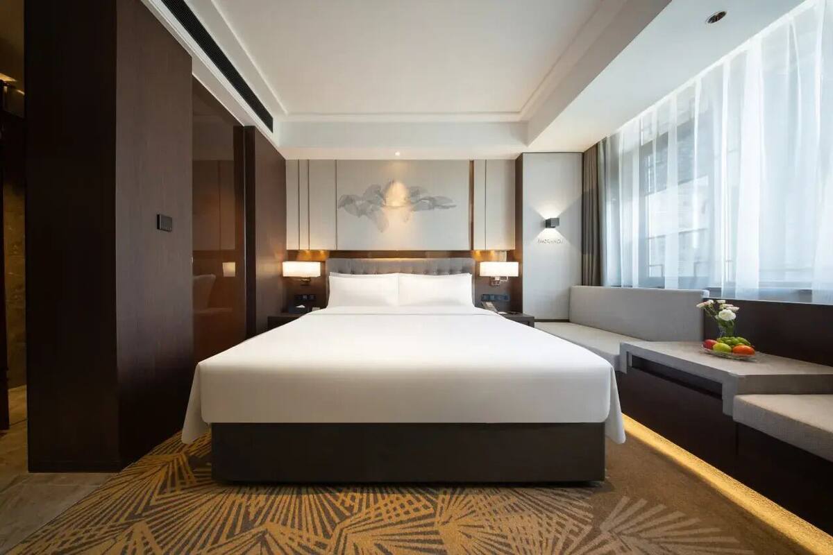 Ein modernes Hotelzimmer mit einem großen Bett, Nachttischen inklusive Lampen, einem Sofa und einem großen Fenster mit leichtem Vorhang.