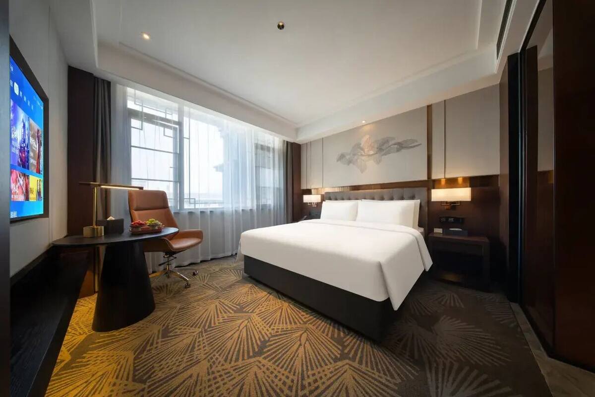 Ein Hotelzimmer mit einem großen Bett, einem Schreibtisch mit Stuhl, einem Fernseher und einem Fenster mit Vorhängen.