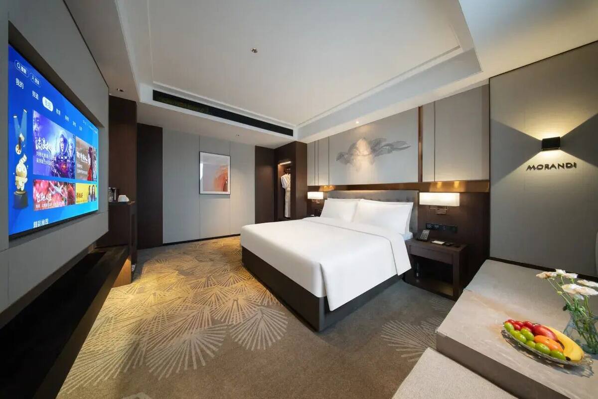 Ein Hotelzimmer mit einem großen Bett, einem flachbildfernseher an der Wand, einem Nachttisch und einem kleinen Tisch mit Obst und Blumen.
