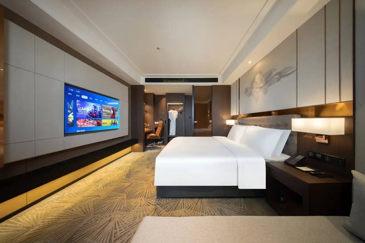 Ein modernes Hotelzimmer mit einem großen Bett, einem an der Wand befestigten Fernseher und einem Schreibtisch mit Lampe.