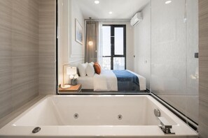 A modern hotel room with a large bathtub, a neatly made bed, a bedside table with a lamp, and a view of the city through the window. - Hampton Residence Thonglor (Bangkok)