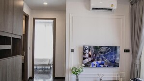 Ett modernt hotellrum med en stor platt-TV som är monterad på väggen, ett träskåp, ett litet bord med en vas med blommor och en säng med en vit sänggavel.