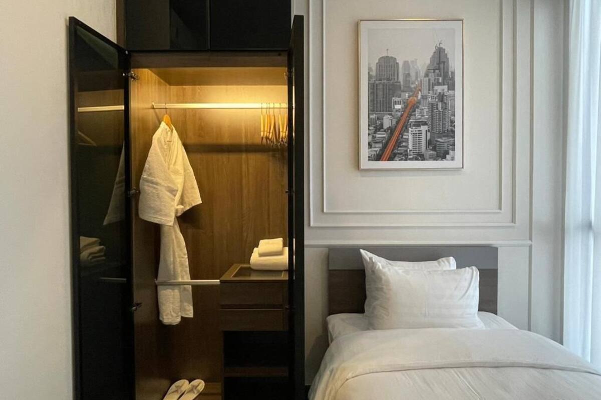 A hotel room with a bed, a wardrobe with a mirror, a framed cityscape picture, and a small table with a towel and slippers.