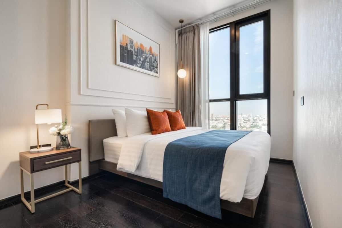 A modern hotel room with a large bed, a bedside table with a lamp, and a cityscape view through the window.
