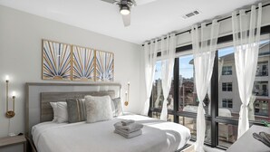 Une chambre moderne avec un grand lit, un ventilateur de plafond et une vue sur la ville à travers des rideaux légers.