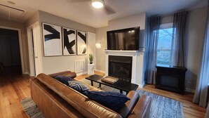 En moderne stue med en lædersofa, et fladskærms-tv, en pejs og et stort vindue med gardiner.