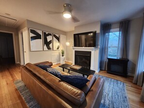 A modern living room with a leather sofa, a flat-screen TV, a fireplace, and a large window with curtains. - Forsyth Park Place, Unit C - Sleeps 4, Three Minute Walk to Forsyth Park (Savannah)