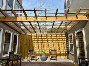 Une structure de pergola en bois, avec un pot de fleurs placé au centre, des chaises en bois et un sol en briques.