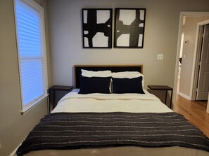 A bedroom with a bed, two nightstands, a window with blinds, and abstract wall art. - Forsyth Park Place, Unit C - Sleeps 4, Three Minute Walk to Forsyth Park (Savannah)