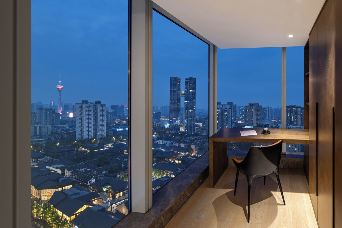 Una habitación moderna con un ventanal grande que ofrece vistas a la ciudad al anochecer, un escritorio con una silla y una mesita con una taza.