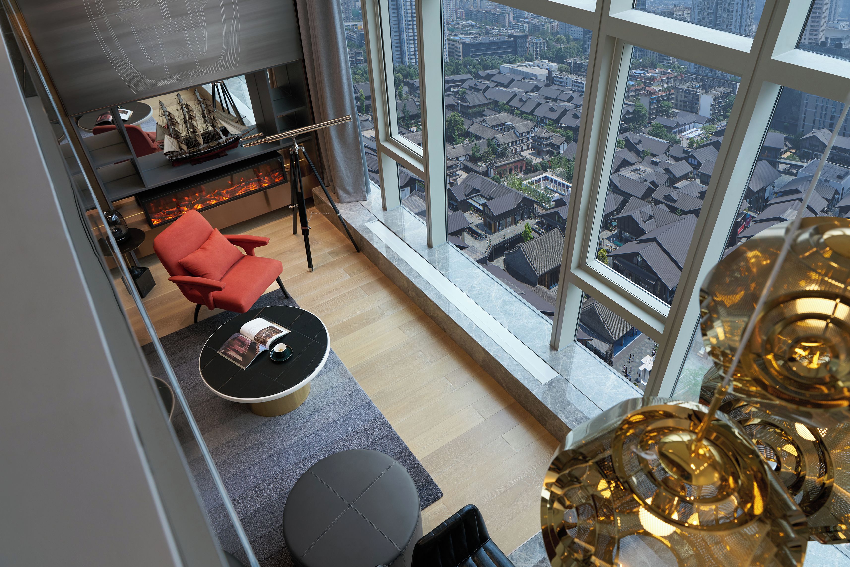 A modern living room with a large window, a red chair, a small round table, and a ship model on a shelf.