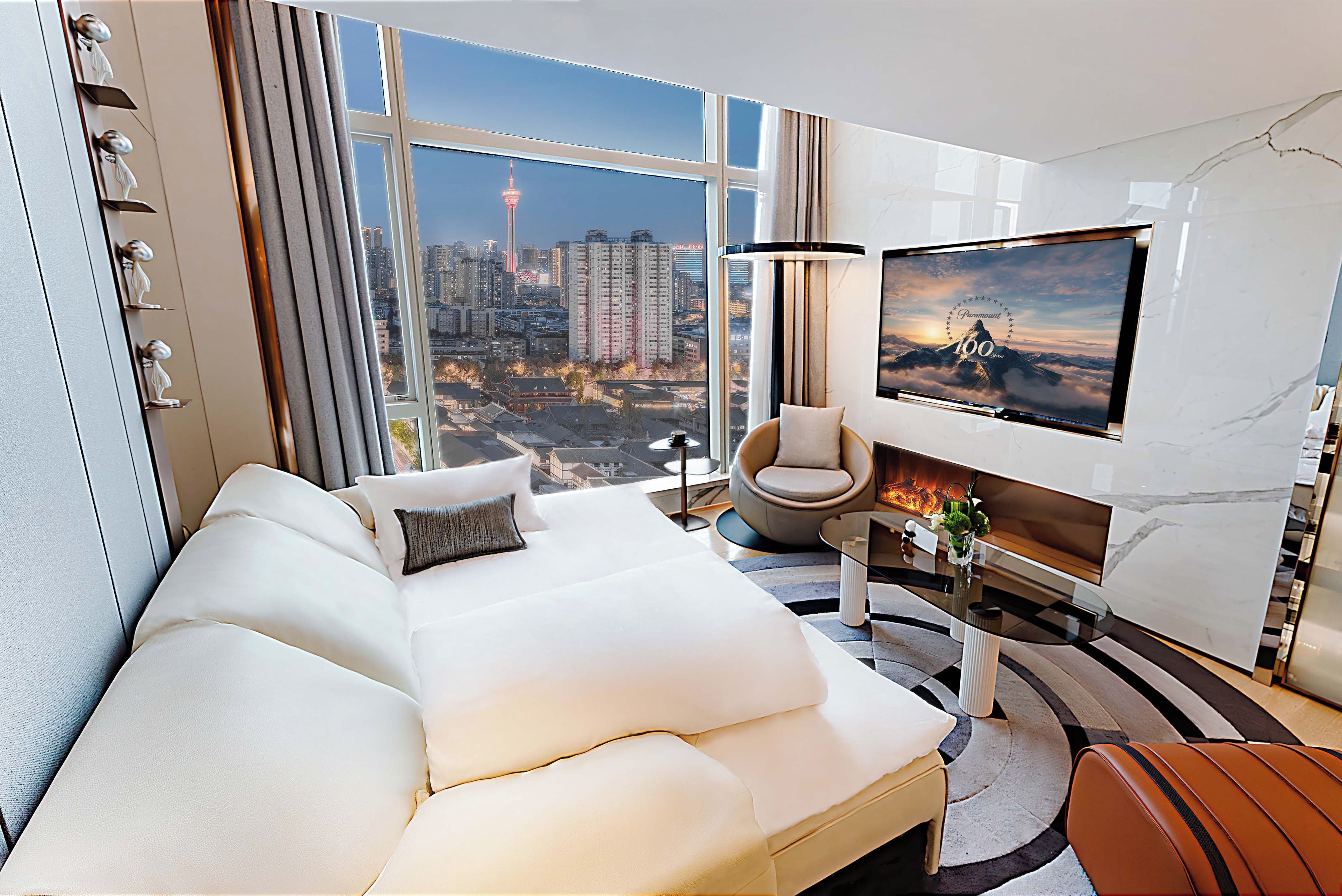 Une chambre d’hôtel moderne dotée d’un grand lit, d’un téléviseur fixé au mur, d’une vue sur le paysage urbain et d’une cheminée.