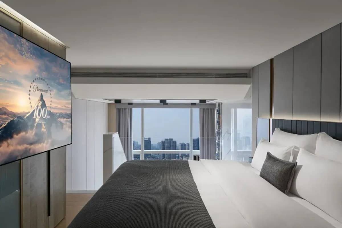 Una camera d'albergo moderna con un letto grande, vista sulla città tramite finestre a tutta altezza e una TV a schermo piatto fissata al muro.