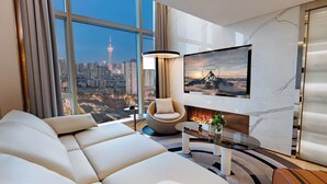 Ein modernes Wohnzimmer mit einer großen Couch, einem runden Couchtisch und einem an der Wand montierten Fernseher. Durch das Fenster ist eine Stadtansicht zu sehen.