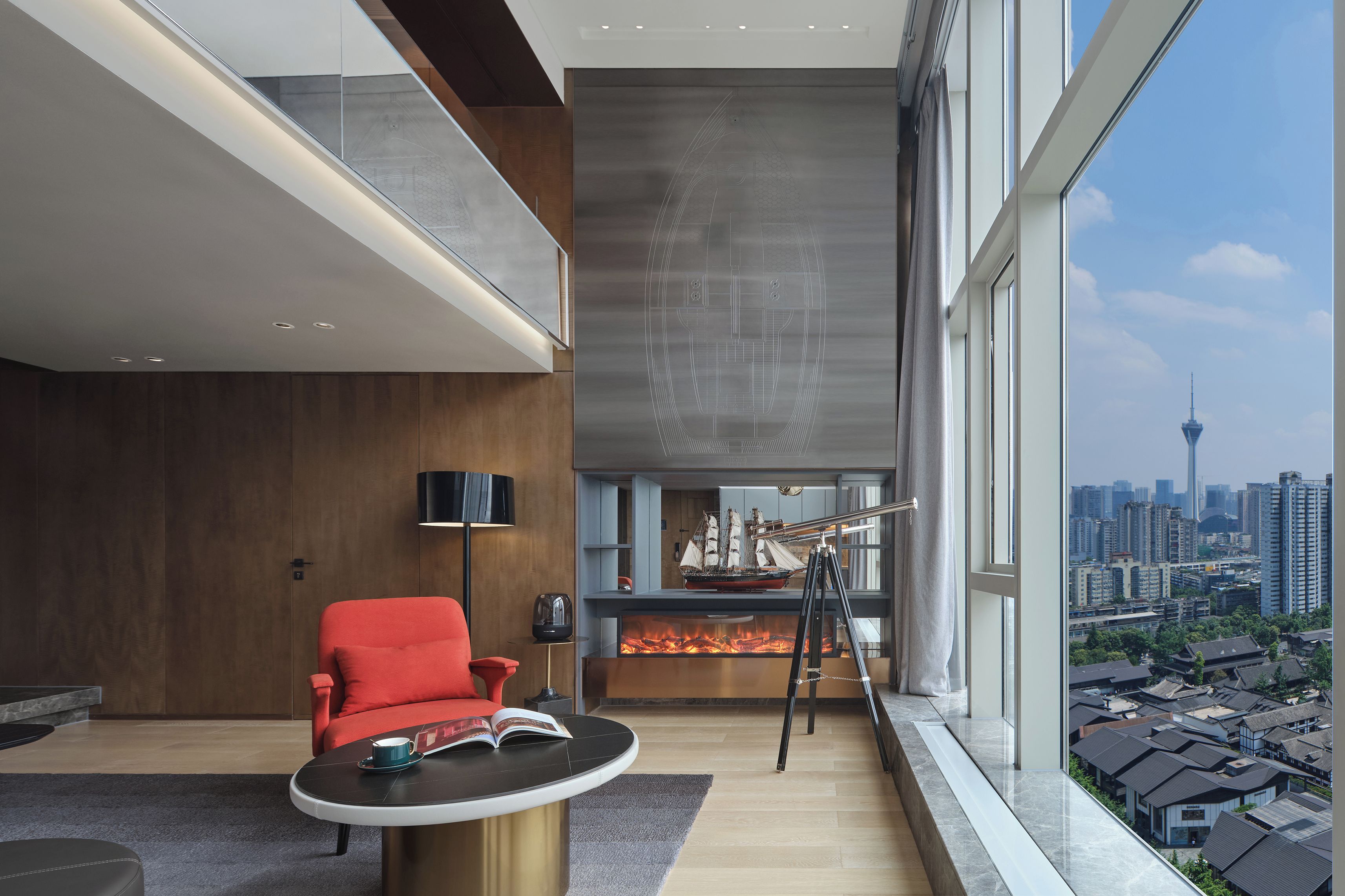 A modern living room with a large window, a fireplace, a red chair, and a table with a book and a cup.