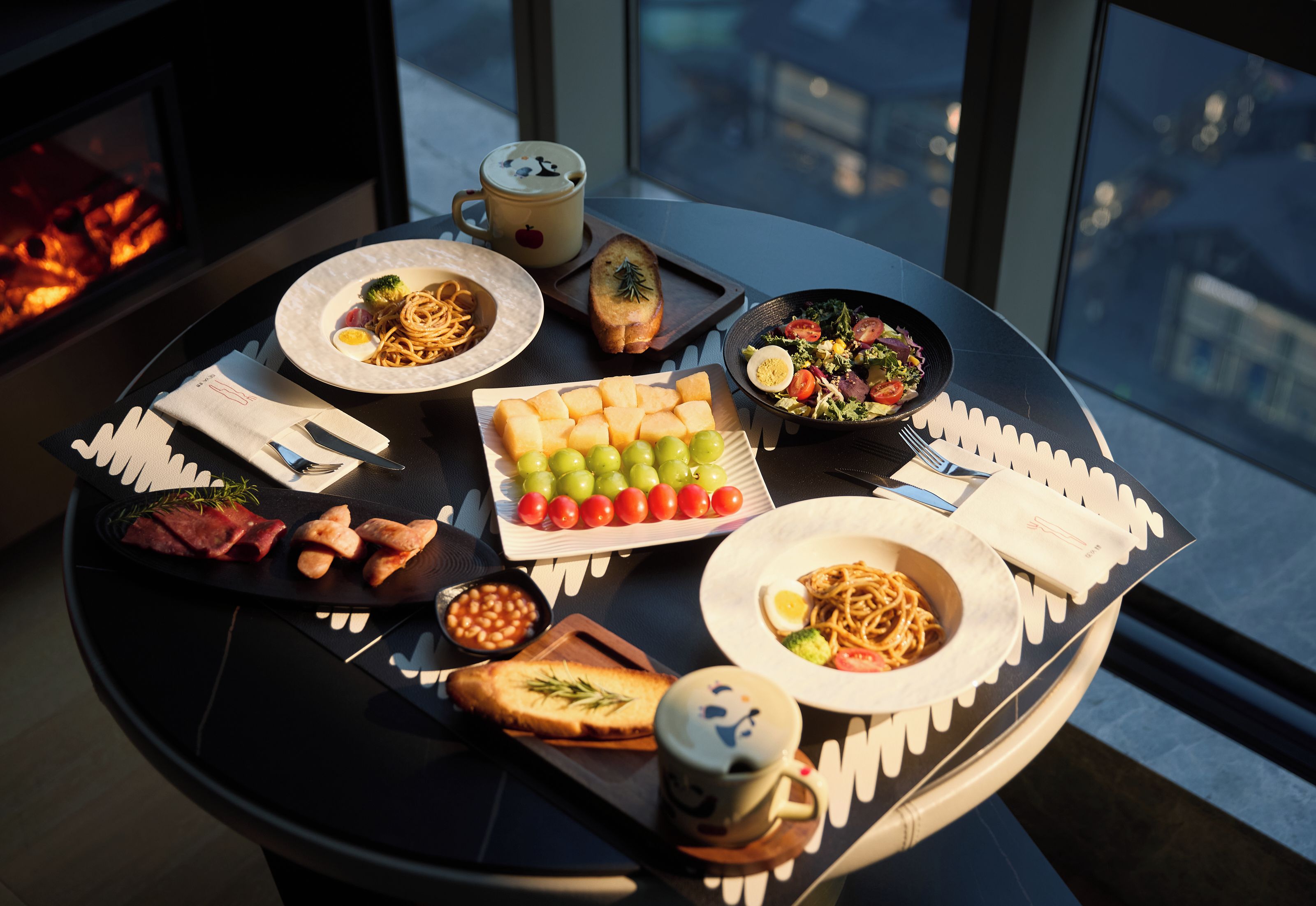 Une table dressée avec divers plats, notamment des pâtes, une salade et du pain, dans une pièce offrant une vue sur les bâtiments à travers de grandes fenêtres.