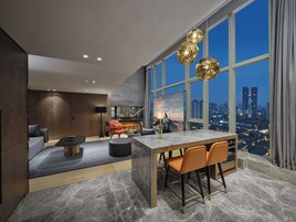 A modern living room with a large window, a dining area, and a view of the cityscape at dusk.