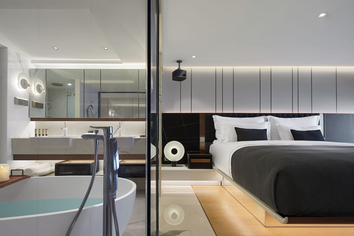 Camera d'albergo moderna con una grande vasca da bagno, una cabina doccia separata, un letto con testiera scura e uno specchio ampio.
