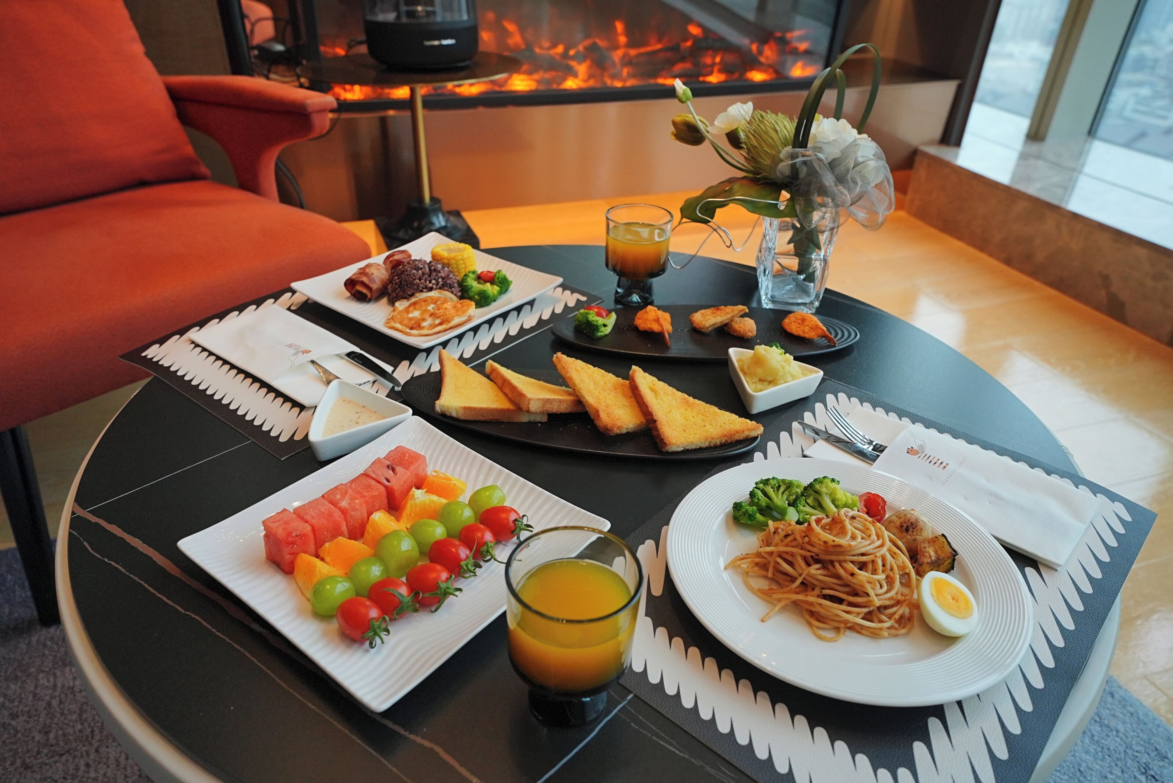 A dining table with a variety of food items, including fruits, vegetables, and a hot beverage, set against a backdrop of a fireplace.