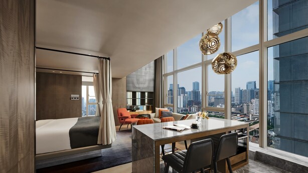 Ein modernes Hotelzimmer mit einem großen Bett, einem Essbereich und Blick auf die Stadt durch große Fenster.