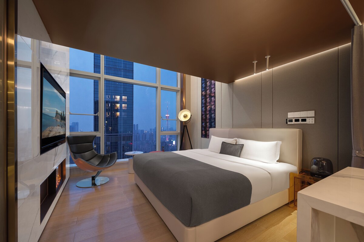 Una habitación de hotel moderna con una cama grande, una zona de estar con una silla, un televisor y vistas a la ciudad.