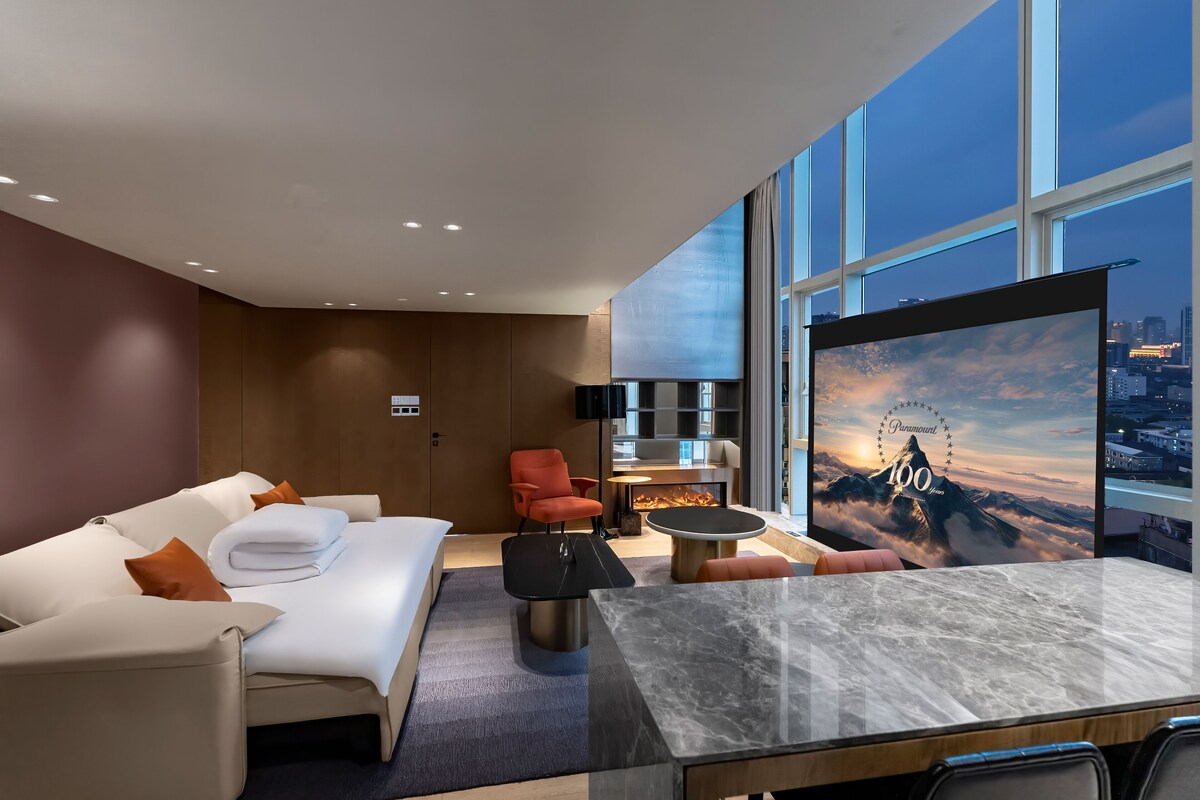 Una habitación de hotel moderna con una cama grande, una zona de estar con una mesita de café y un televisor que muestra una vista de la montaña.