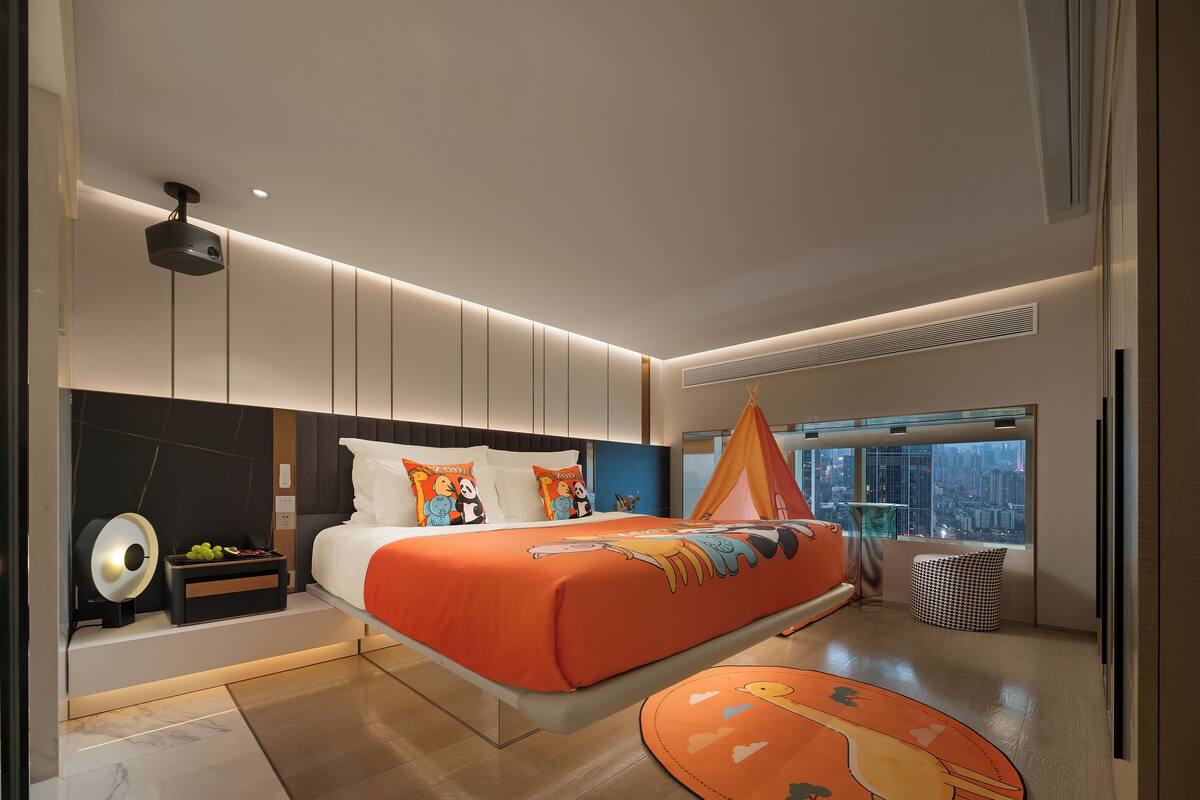Una camera da letto moderna con un letto grande, un comodino con della frutta, un proiettore a parete e vista sulla città attraverso un'ampia finestra.