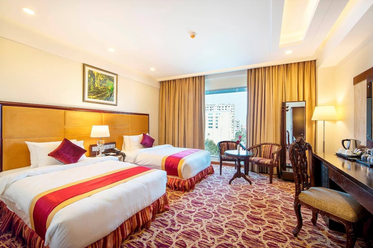 Quarto de hotel com duas camas, uma escrivaninha, uma cadeira e uma janela grande com cortinas.