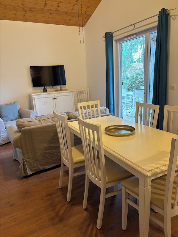 A dining area with a white table and chairs, a television above the table, and a view of the outdoors through a window with blue curtains.