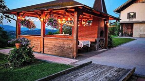 A wooden gazebo with a brick chimney, surrounded by a grassy area and a paved path leading to a building. - Charming family apartment with terrace, herb garden views and animals (Štore)