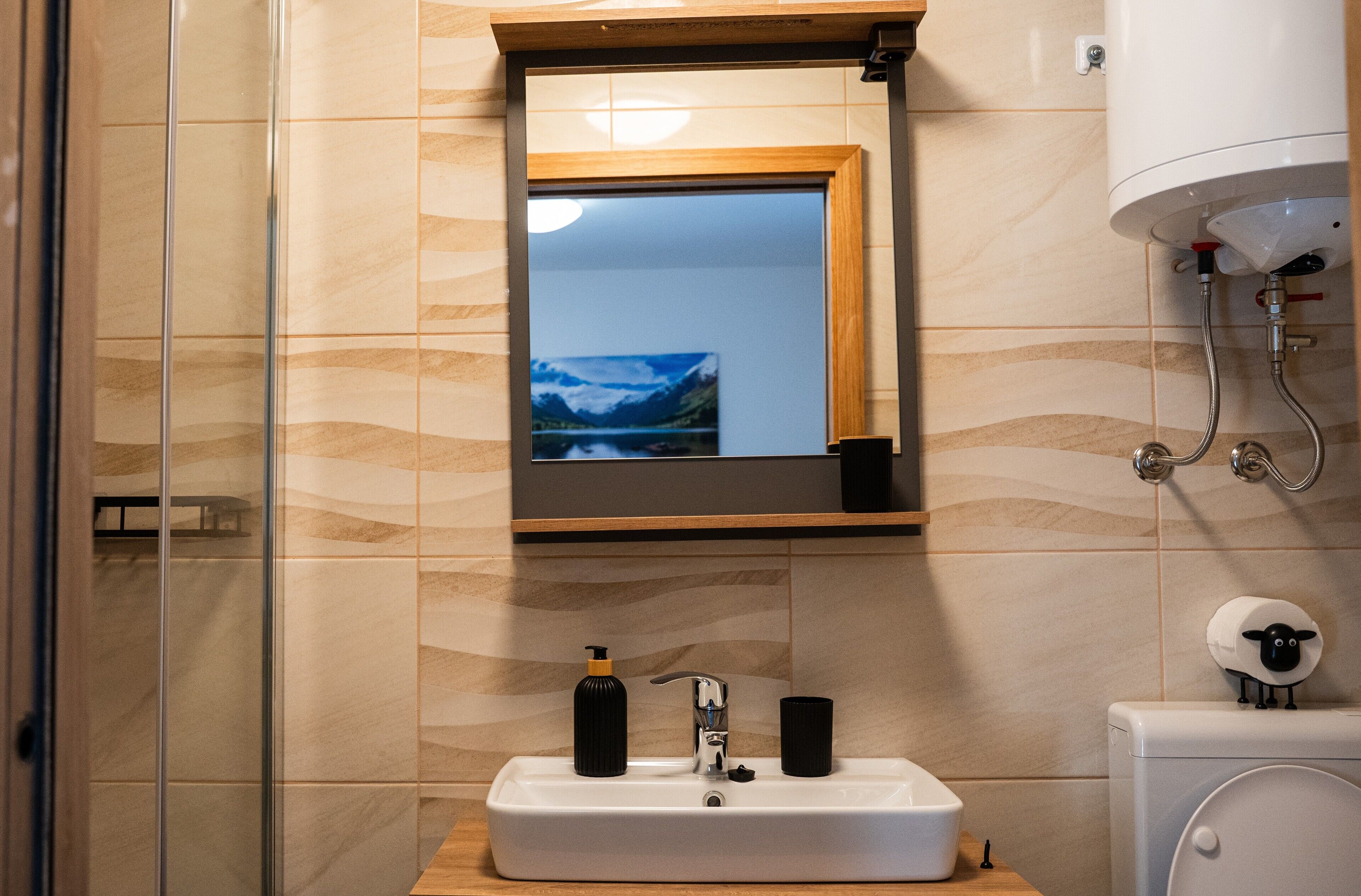 Un baño con un lavabo blanco, dos dispensadores de jabón negros, un espejo sobre el lavabo y un inodoro decorado con un motivo de oveja.