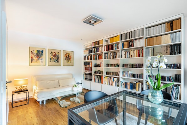 Un salon moderne avec un canapé blanc, une table basse en verre, une bibliothèque remplie de livres et un vase contenant des orchidées blanches.