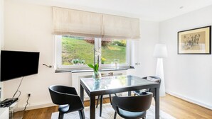 A modern room with a TV, a glass table, black chairs, a window with a view of greenery, and a framed picture on the wall. - Aachen South Holiday Apartment for 4 Guests (55 m²) (Aachen)
