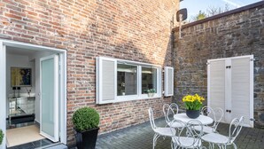 A brick building with a patio area featuring a white metal table and chairs, a potted plant, and a door leading to an interior space. - Aachen South Holiday Apartment for 4 Guests (55 m²) (Aachen)