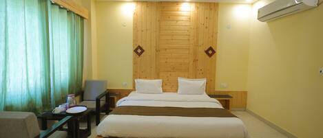 A hotel room with a wooden headboard, a bed with white bedding, a small table with a chair, and a window with green curtains.