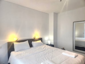 En pænt redt seng med hvide sengetøj, to sengeborde med lamper, og et stort spejl på væggen.