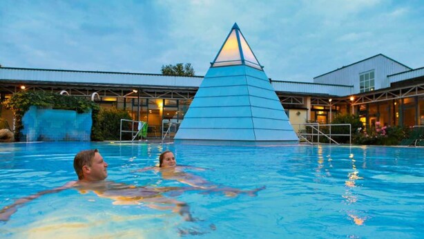 Ein Schwimmbecken mit einer pyramidenförmigen Struktur in der Mitte, umgeben von einem Gebäude mit Metallbedachung und Glasfenstern.