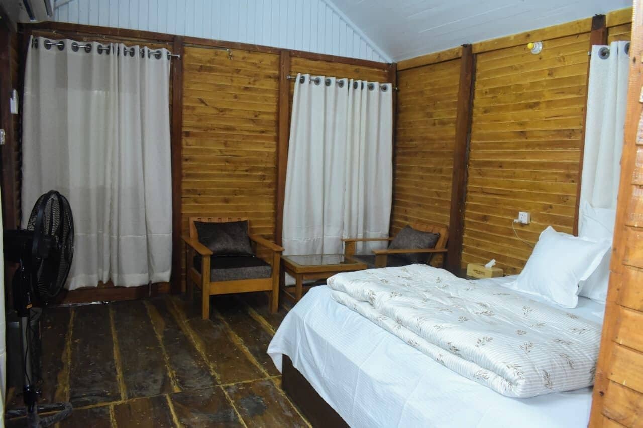 Quarto com uma cama, duas cadeiras, uma mesinha e um ventilador.