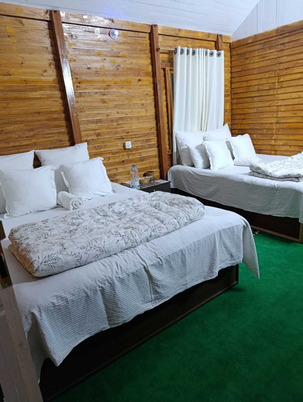 A room with two beds, wooden walls, a green carpet, and a white curtain.