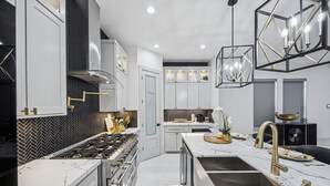 A modern kitchen with white cabinetry, a black herringbone backsplash, a gas stove, and a large island with a sink and gold faucet. - One Story + Generator & Gated Near HTX Medical (Houston)