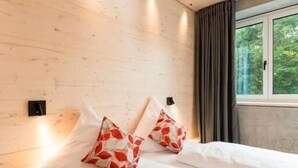 A bedroom with a wooden ceiling, two beds with white bedding, red and white patterned pillows, a window with a view of greenery, and a nightstand with a book. - Hohentwiel Apartments (Singen)