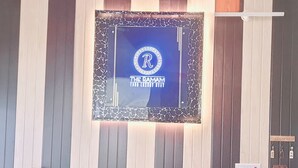 En man sitter i ett rum. På väggen hänger en inramad logotyp, och i rummet finns ett litet bord med en dekorativ prydnad samt en väggmonterad elcentral.
