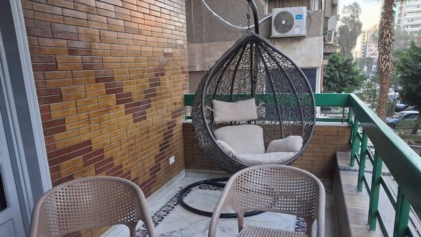 A balcony with two wicker chairs, a hanging chair with cushions, and a green railing. - Spacious 3-Bedroom Apartment in Central Cairo with City View (Cairo Governorate)