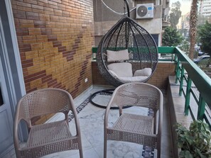 Un balcón con dos sillones de mimbre, un sillón colgante con cojines y una barandilla verde.