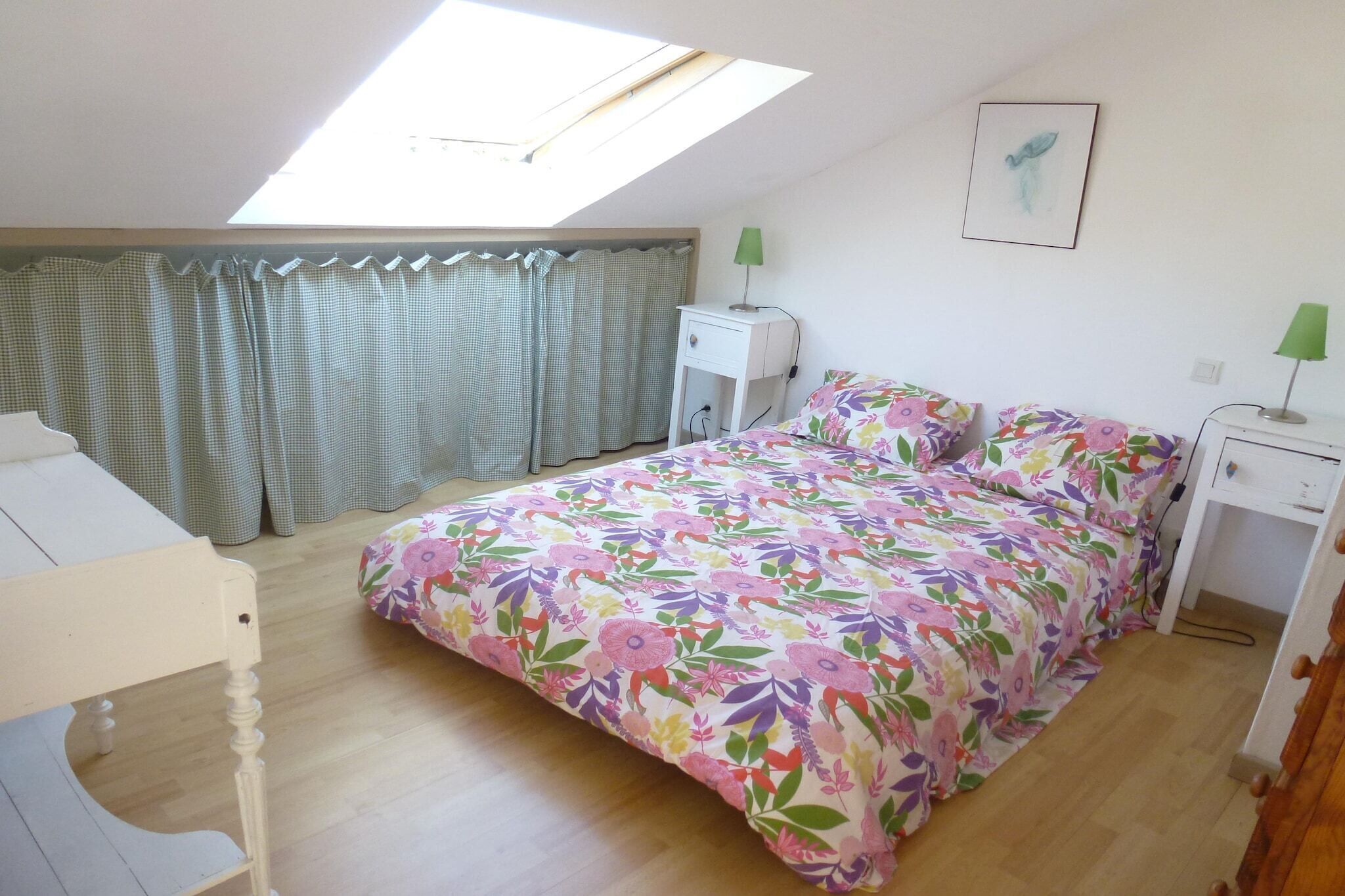 A bedroom with a skylight, a bed with floral bedding, a bedside table with a lamp, and a desk with a chair.