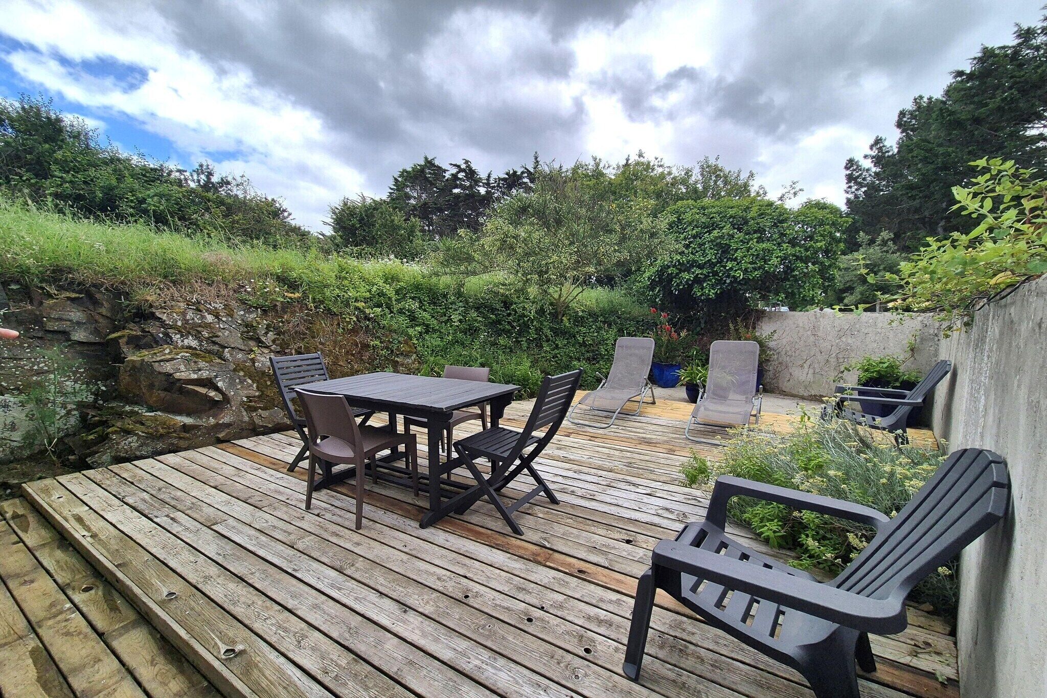 Terraza de madera con mesa y sillas, rodeada de follaje y un muro de piedra.