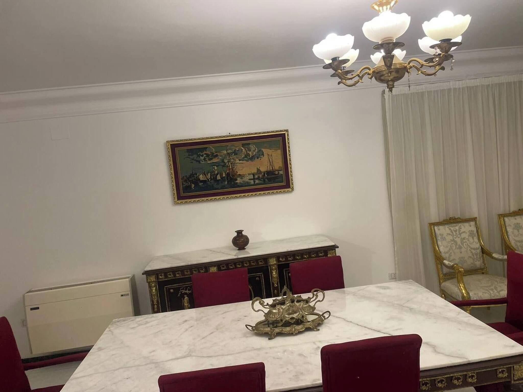 Un comedor con una mesa de mármol, sillas tapizadas en rojo, una araña y un cuadro en la pared.